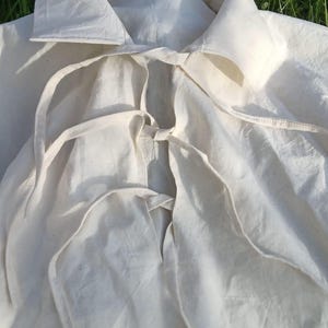 May include: A white cotton garment with a large collar and a tie closure. The garment is folded and laid out on a bed of green grass.
