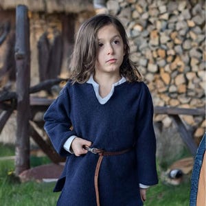 May include: A child wearing a navy blue tunic with a white collar and cuffs, cinched at the waist with a brown leather belt. The tunic is paired with burgundy pants. The background features a wooden fence and a thatched roof.