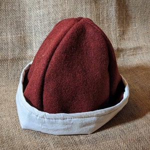 May include: A burgundy felt hat with a white fabric brim. The hat has a rounded crown and a simple design.