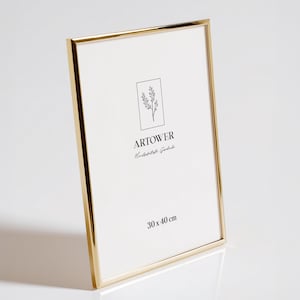 Könnte beinhalten: Ein goldfarbener Metall-Bilderrahmen mit weißem Innenraum. Der Rahmen trägt den Text "ARTOWER" und "30 x 40 cm" auf dem weißen Innenraum. Der Rahmen ist leicht nach links geneigt.