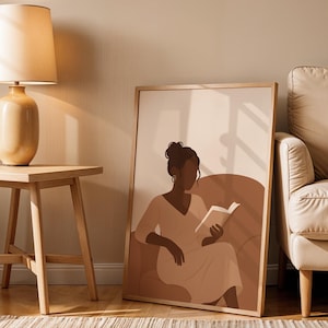 May include: A framed art print featuring a minimalist illustration of a person reading a book while seated. The artwork uses a warm, neutral color palette with shades of brown and beige. The print is displayed next to a wooden side table and a beige sofa.