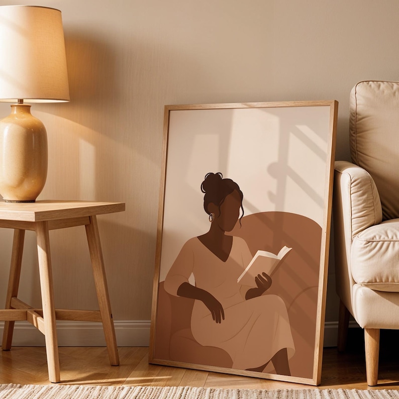 May include: A framed art print featuring a minimalist illustration of a person reading a book while seated. The artwork uses a warm, neutral color palette with shades of brown and beige. The print is displayed next to a wooden side table and a beige sofa.