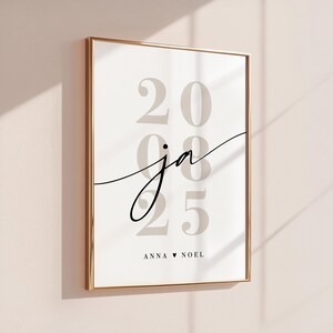 May include: A framed wall art print featuring the large, light gray numbers 2025 stacked vertically. Overlaid in the center is the black cursive text ja. At the bottom, the text ANNA &hearts; NOEL is printed in a simple, clean, black sans-serif font.