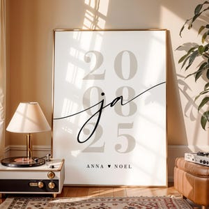 May include: A vertical rectangular wall art print in a gold frame featuring the large light gray numbers 2025. Overlaid in the center is the black cursive script letters ja. Below the numbers, the text ANNA &hearts; NOEL is printed in black.