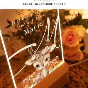May include: A personalized night light with a deer illustration and floral accents. The light features a clear acrylic panel with the name "Maria", the time "20:22", and the weight "3245 g" and height "53 cm". The light has a wooden base.