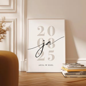 May include: A rectangular framed wall art print featuring the large light gray numbers 20 and 25 stacked vertically. Overlaid in the center is the lowercase script text ja in black. At the bottom, the text ANNA, a heart symbol, and NOEL appear.
