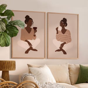 May include: Two framed wall art prints. Each print features a minimalist illustration of a person in a seated pose. One holds a glass of red wine, the other holds a small item. The figures are in neutral tones, with a light beige background and wooden frames.