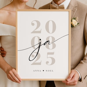 Könnte beinhalten: Ein gerahmter Druck mit den Zahlen 20, 25 und dem Wort "ja" in einer Schreibschrift. Die Namen "ANNA ♥ NOEL" sind unten aufgedruckt. Der Druck wird von zwei Personen gehalten, eine in einem beigen Anzug und die andere in einem weißen Kleid.