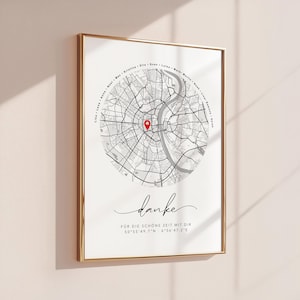 Puede incluir: Impresi&oacute;n enmarcada con un mapa en blanco y negro y un marcador rojo. La palabra "danke" est&aacute; escrita en cursiva debajo del mapa, junto con coordenadas y texto. El marco es dorado.