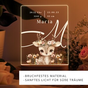 May include: A white acrylic night light with a deer illustration and the text "Maria" and birth details: 20:22 Uhr, 3245 g, 22.08.23, 53 cm. The night light has a wooden base and the text "BRUCHFESTES MATERIAL - SANFTES LICHT F&Uuml;R S&Uuml;BE TR&Auml;UME".