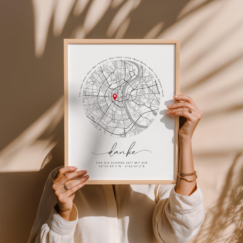 Puede incluir: Una impresi&oacute;n enmarcada con un mapa en blanco y negro y un marcador rojo. La palabra "danke" est&aacute; escrita en cursiva debajo del mapa, junto con las coordenadas. La impresi&oacute;n est&aacute; sostenida por una persona.