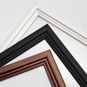 Könnte beinhalten: Nahaufnahme von drei Bilderrahmen in Weiß, Schwarz und Braun. Die Rahmen haben eine abgeschrägte Kante und sind zum Betrachter hin abgewinkelt. Der weiße Rahmen hat eine subtile Holzmaserung, während die schwarzen und braunen Rahmen eine glatte Oberfläche haben.