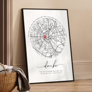 Puede incluir: Impresi&oacute;n enmarcada de un mapa en blanco y negro con un marcador rojo. El mapa es de una ciudad, con nombres en c&iacute;rculo en la parte superior. La palabra "danke" est&aacute; escrita en cursiva debajo, con coordenadas.