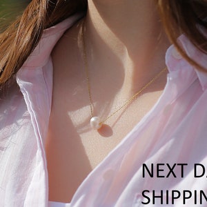 May include: A delicate gold chain necklace with a single white pearl pendant. The necklace is worn by a person with long brown hair and a pink shirt. The text "NEXT DAY SHIPPING" is visible at the bottom of the image.