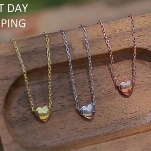May include: Three delicate gold, silver, and rose gold necklaces with small heart pendants. The necklaces are displayed on a wooden surface.
