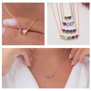 May include: A gold necklace with a row of five small heart-shaped charms, each with a different coloured gemstone. The gemstones are green, pink, blue, white and red.
