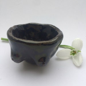 May include: A small, dark brown ceramic bowl with a textured, uneven surface. The bowl has a rounded shape and a slightly flared rim. It is sitting on a white surface.