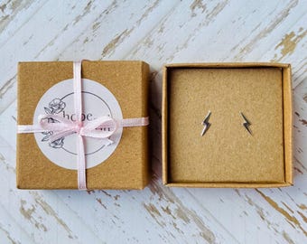Puede incluir: Un par de pendientes de botón de rayo plateado en una caja de cartón marrón con una cinta rosa y una pegatina blanca con el texto "hope & whim".