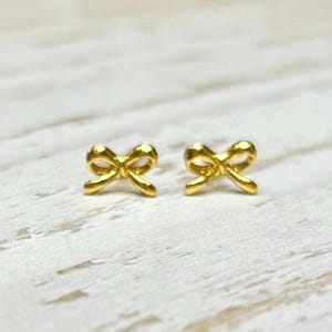 Pode incluir: Um par de brincos pequenos dourados em forma de laço. Os brincos estão colocados numa superfície clara e texturizada. Os laços têm uma forma arredondada e um acabamento brilhante.
