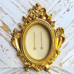 Könnte beinhalten: Ein Paar goldene herzförmige Ohrringe mit einer zarten Kette, die von jedem Herzen hängt. Die Ohrringe werden in einem goldenen, verzierten ovalen Rahmen präsentiert.