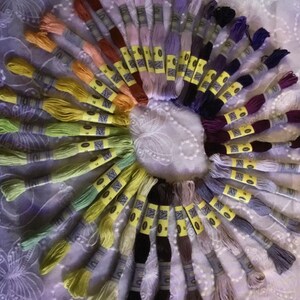 May include: A collection of embroidery floss spools arranged in a circle, showcasing a variety of colors, including shades of green, yellow, purple, blue, and red. Each spool has a yellow label with a number and a barcode.