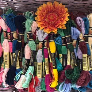 May include: A selection of vibrant embroidery threads in various colours, such as pink, blue, green, and yellow. An orange gerbera daisy rests on top of the threads. The threads are neatly arranged, indicating a craft or hobby supply.