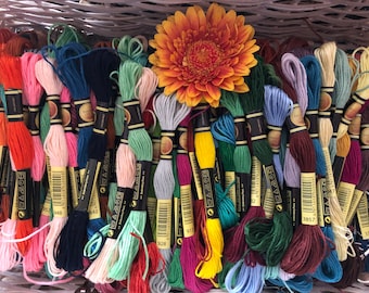 May include: A collection of colorful embroidery floss in a variety of shades, including pink, blue, green, and yellow. A vibrant orange gerbera daisy sits atop the floss. The floss is neatly arranged, suggesting a craft or hobby supply.