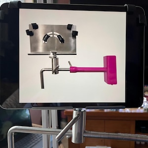 May include: A silver-coloured metal device with a pink handle and a rectangular head. The device is mounted on a tablet, with black straps securing it. The device has a complex design with multiple joints and a curved section.