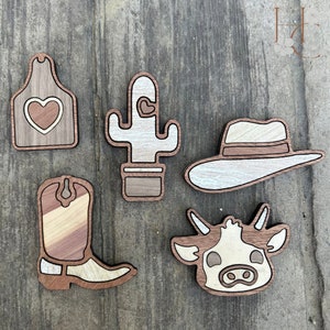 May include: Five wooden cutouts in a rustic theme. The shapes include a tag with a heart, a cactus, a cowboy hat, a cowboy boot, and a cow's head. The cutouts are made of layered wood in shades of brown, tan, and white.
