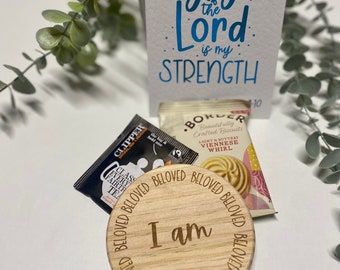 May include: A wooden round coaster with the words "I am" in the center and the word "BELOVED" repeated around the edge. The coaster is sitting on a white surface next to a package of tea and a package of cookies. The text on the card reads "The joy of the Lord is my STRENGTH".