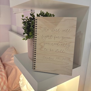 May include: A wooden notebook with a spiral binding, inscribed with the words "The Lord will fight for you, you need only to be still Exodus 14. 14." It sits on a white shelf, next to a small potted plant. The background includes a bed and a wall.