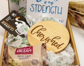 May include: A gift box filled with items including a wooden round plaque with the word "Cherished" engraved on it, a tea bag, a bag of chocolate chip biscuits, and a card with the text "Love is my strength".