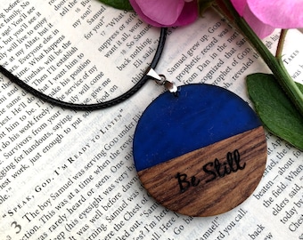 May include: A wooden pendant necklace with a blue and brown circular design. The pendant has the words "Be Still" engraved on the brown wood. The necklace is on a black cord.