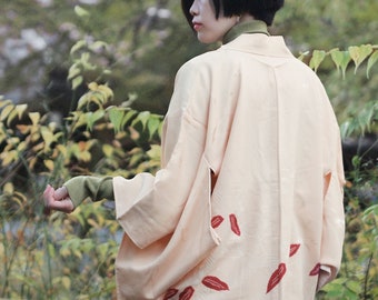 Puede incluir: Una mujer lleva un kimono de color melocotón pálido con bordados de hojas rojas. El kimono tiene mangas anchas y un corte holgado. La mujer está de pie en un jardín con follaje verde.
