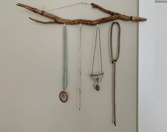 May include: A driftwood branch mounted on a wall with four necklaces hanging from it. The necklaces are made of different materials, including beads, metal, and leather.
