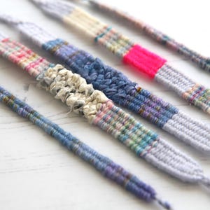 May include: Close-up of several handmade friendship bracelets. Each bracelet features a unique design with various colours, including blue, pink, and white. The bracelets are made of woven fabric and are displayed on a white surface.