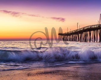 May include: A wooden pier extends out over a calm ocean at sunrise. The sky is a vibrant mix of pink, orange, and purple. A wave crashes on the sandy beach in the foreground.