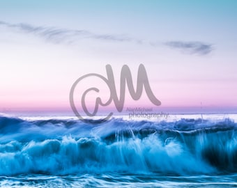 May include: A large wave crashes against a wooden pier at sunset. The sky is a gradient of pink, purple, and blue. The water is a deep blue with white foam.