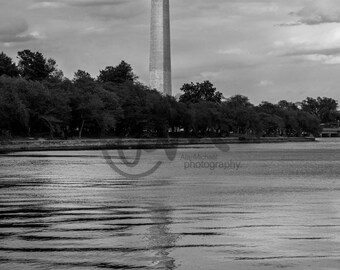 May include: A black and white photograph of the Washington Monument in Washington, D.C. The monument is tall and slender, with a pointed top. It is surrounded by trees and water. The sky is cloudy.