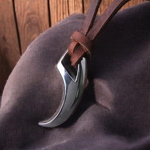 May include: A polished silver pendant in the shape of a claw, suspended from a brown leather cord. The claw pendant has a curved, pointed design and is attached to the cord with a loop. The background is a dark, textured fabric.