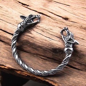 May include: A silver bracelet with a wolf head design on each end. The bracelet is made of twisted metal and has a detailed design.