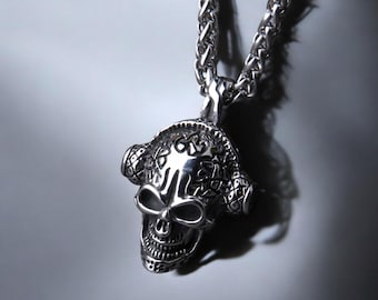 May include: A silver-colored skull pendant necklace. The skull has detailed features, including eye sockets, a nose, and teeth. The pendant is attached to a chain with a textured, woven design. The necklace is displayed against a gray background.