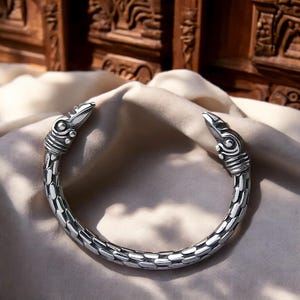 May include: A silver bracelet with a detailed design of two bird heads on the ends. The bracelet is made of a textured metal and has a rounded shape.