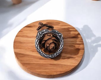 May include: A silver ring with a braided design and a textured, raised design on the top. The ring is on a wooden surface.