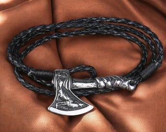 May include: A black braided leather bracelet with a silver-colored axe pendant. The axe features intricate engraved details. The bracelet is wrapped around itself, showcasing the design against a brown background.