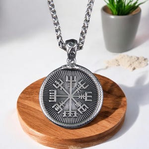 May include: A silver-colored pendant necklace featuring a circular pendant with a detailed Viking compass design. The pendant is suspended from a thick, textured chain. The necklace is displayed on a wooden surface.