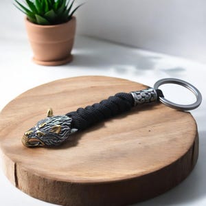 May include: A black paracord keychain with a silver wolf head charm and a silver ring. The wolf head has gold accents.