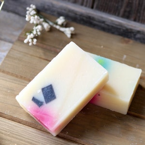 Op de afbeelding: Twee handgemaakte zeepjes, een met een roze en witte swirl en een zwart diamantontwerp, de andere met een blauwe en witte swirl. De zeepjes liggen op een houten oppervlak.