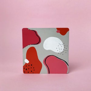 May include: A square coaster with an abstract design featuring pink, red, white, and black shapes on a gray background.