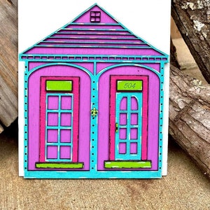 May include: A wooden cutout of a pink house with blue trim and green accents. The house has two doors and a window. The number 504 is written above one of the doors.